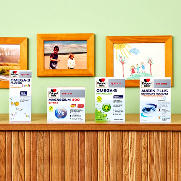 Produktaufsteller mit vier verschiedenen Nahrungsergänzungsmitteln von Doppelherz. Die Produkte sind Magnesium 400 Citrate, Omega-3 Plüssig, Omega-3 Pflanzlich und Augen Plus. Im Hintergrund sind familienfreundliche Bilder in einem hellen Raum angebracht.