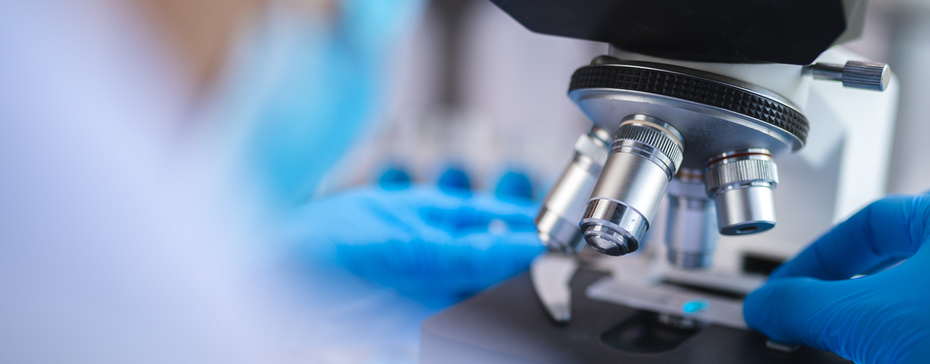 Das Bild zeigt einen Mikroskop, der von jemandem mit blauen Handschuhen bedient wird. Im Hintergrund sind unscharfe labortechnische Geräte sichtbar. Das Hauptaugenmerk liegt auf der Präparatsheibe unter dem Mikroskop.