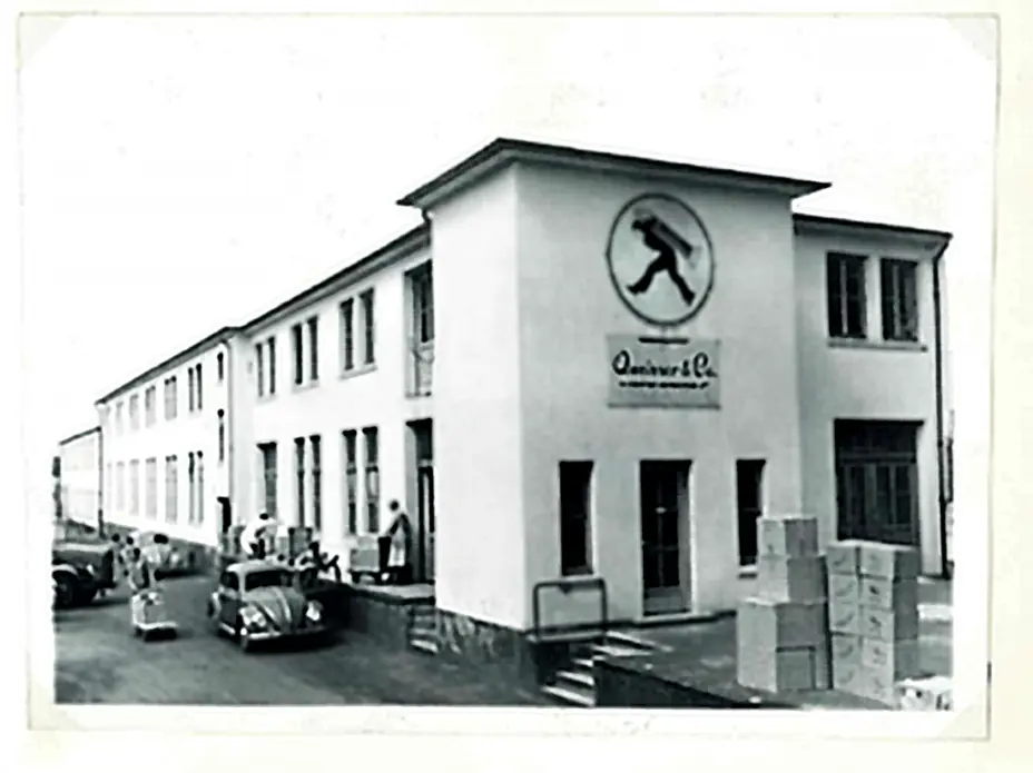 Ein historisches schwarz-weiß Foto eines Gebäudes mit einem großen Schild, das eine stilisierte Figur zeigt. Vor dem Gebäude stehen einige Personen und Autos, während an der Seite Kisten gestapelt sind. Das Gebäude hat große Fenster und eine Eingangstür.