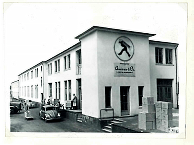 Une vue en noir et blanc d'un bâtiment commercial avec un logo sur la façade. Des personnes et des voitures anciennes sont visibles à l'extérieur. Des caisses sont empilées sur le côté. L'atmosphère suggère une époque révolue, probablement les années 1950.