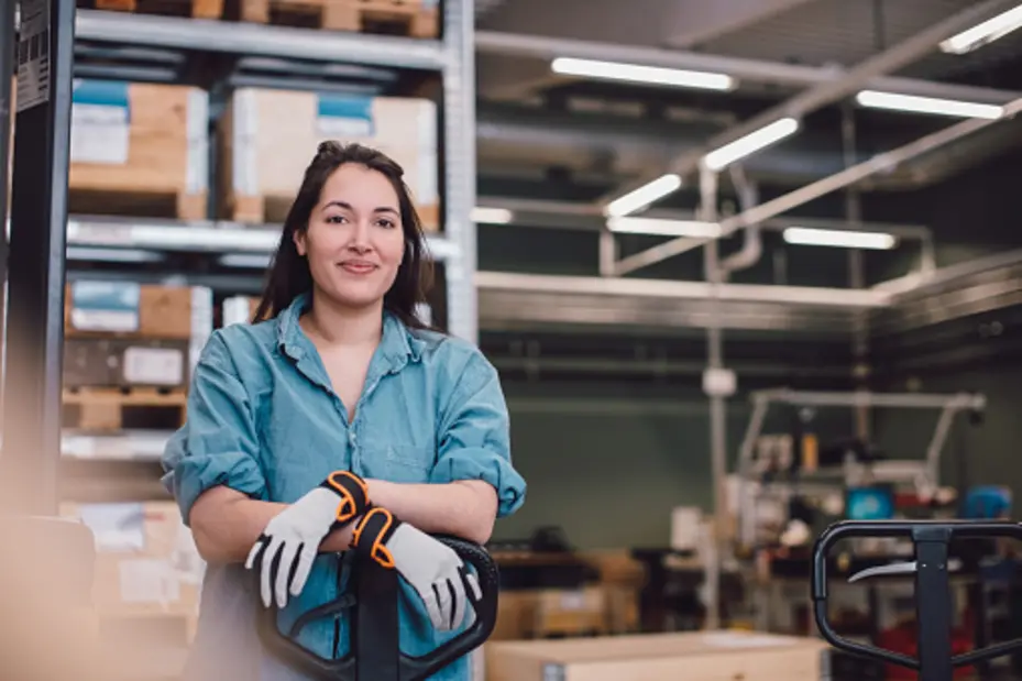 Eine arbeitende Frau in einem Lager steht am Gabelstapler. Sie trägt eine blaue Bluse und Arbeitshandschuhe. Im Hintergrund sind Regale mit Lagerboxen zu sehen, was auf eine industrielle Umgebung hinweist.