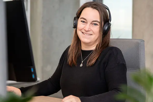 Eine Person mit Kopfhörern sitzt an einem Schreibtisch vor einem Computer und lächelt. Der Hintergrund zeigt einen modernen Arbeitsraum mit hellem Licht.