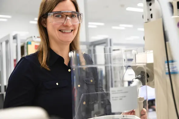 Eine lächelnde Frau trägt Schutzbrille und steht neben einem Laborgerät. Im Hintergrund sind Regale mit wissenschaftlichen Materialien sichtbar. Die Atmosphäre wirkt freundlich und professionell.