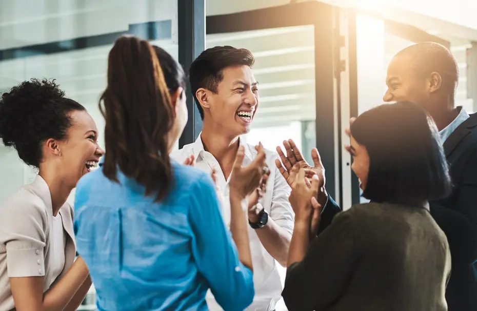 Eine Gruppe von fünf Personen steht in einem modernen Raum. Sie lachen und applaudieren einander, was auf eine fröhliche, unterstützende Atmosphäre hindeutet. Die Personen sind vielfältig in Aussehen und tragen legere Kleidung.