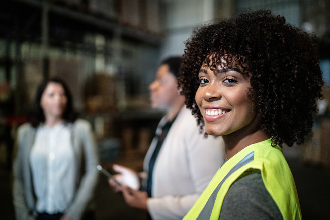 Une femme souriante avec des cheveux bouclés porte un gilet de sécurité. Elle se tient au premier plan, tandis que deux autres femmes, en arrière-plan, discuttent. L'environnement semble être un entrepôt ou un espace de travail.