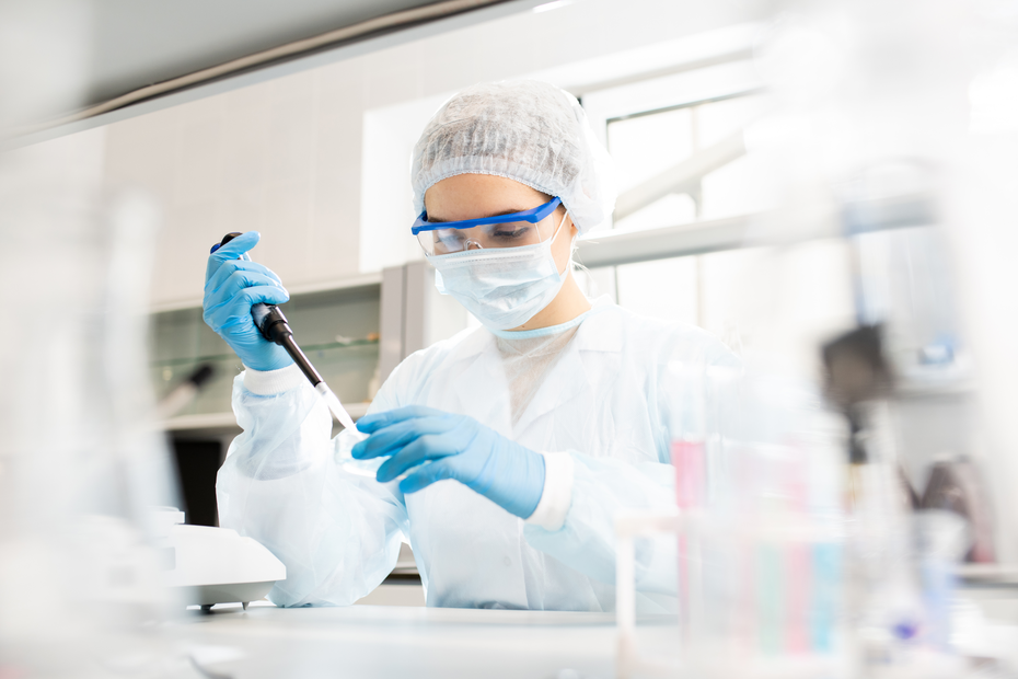 Eine Laborantin in Schutzkleidung, mit Handschuhen und einer Schutzmaske, verwendet eine Pipette, um eine Flüssigkeit in einem Labor zu transportieren. Um sie herum sind verschiedene Laborausrüstungen sichtbar.