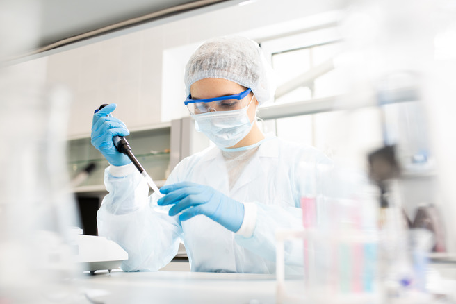 Une scientifique en blouse blanche, portant un masque et des gants, utilise un pipette dans un laboratoire. Elle est concentrée sur son travail, avec des équipements de laboratoire visibles en arrière-plan.