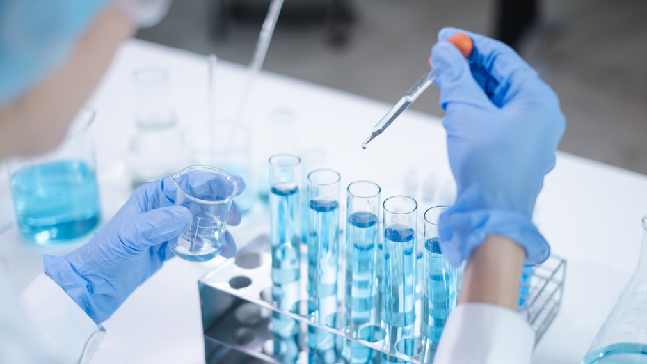 Eine Person in Schutzkleidung hält ein Becherglas mit einer blauen Flüssigkeit in der linken Hand und eine Pipette in der rechten Hand. Vor ihr stehen mehrere Reagenzgläser, die ebenfalls mit blauer Flüssigkeit gefüllt sind.
