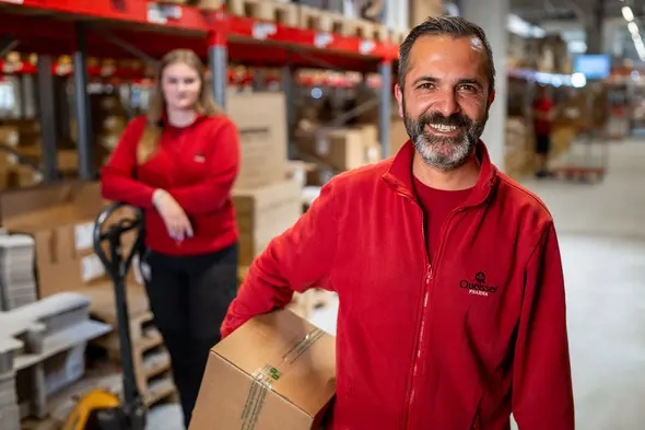 Ein fröhlicher Mann trägt ein Paket und steht in einem Lager, während eine Frau im Hintergrund in rotem Oberteil neben einem Handhubwagen steht. Beide scheinen in einem geschäftigen Arbeitsumfeld zu sein, umgeben von Kartons.
