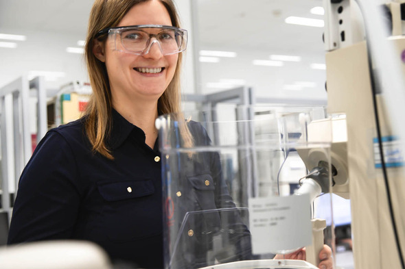 Femme souriante portant des lunettes de protection dans un laboratoire. Elle est devant un équipement scientifique, avec des étagères et des appareils en arrière-plan. L'image dégage une ambiance de travail scientifique et de sécurité.